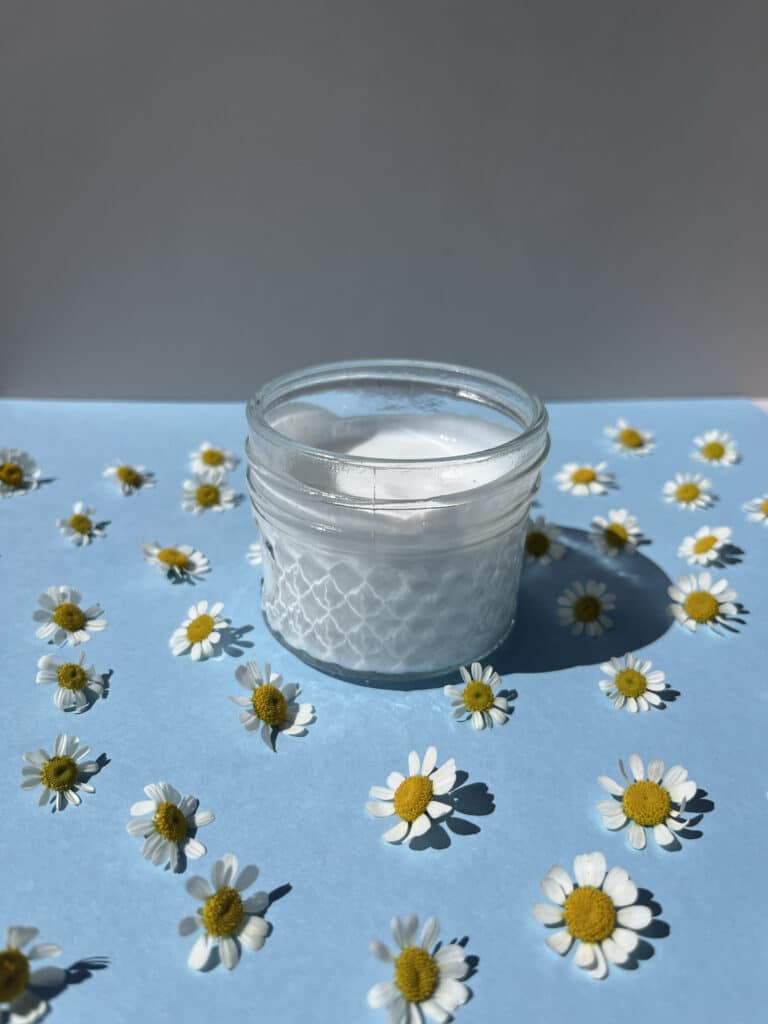 Glass jar with moisturizer surrounded by daisy flowers on a blue background.
