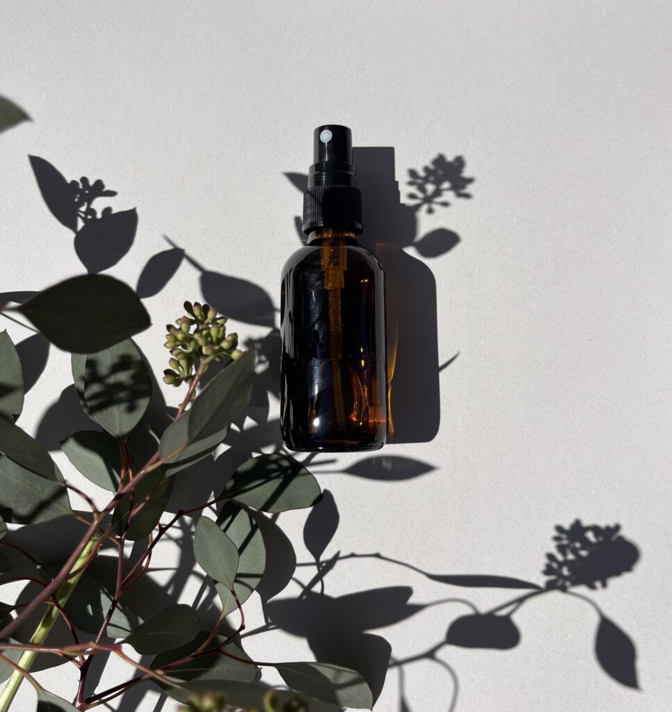 Amber color glass spray bottle surrounded by leaves and shadows on a white background.
